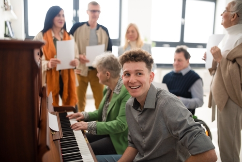 A young man sits at a piano with an older woman. Several people watch from the background.