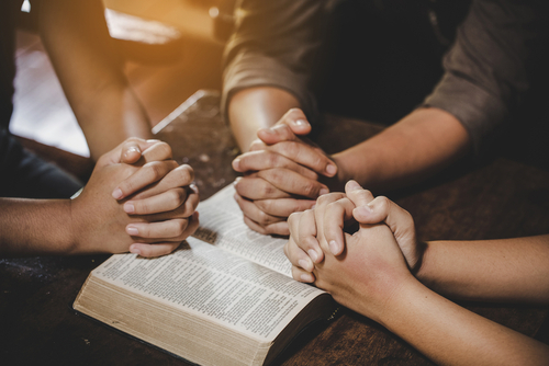 Three people fold their hands over a bible and pray together.