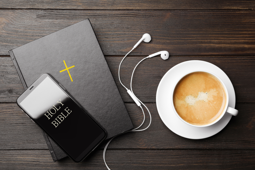 A phone with earbuds sits on top of a Bible and next to a cup of coffee.