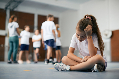 A young school girl its alone while other kids play in the background.