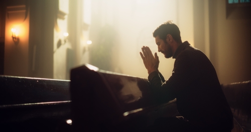 A man sits and prays alone on the pew of a darkened church.