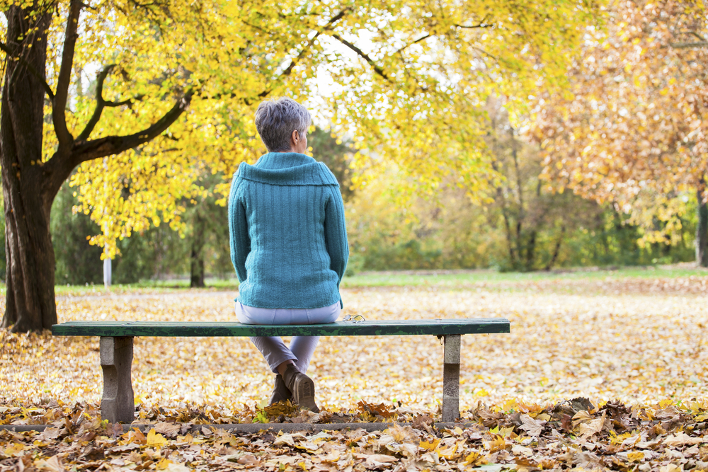 An older woman sits alone on a park bench with fall colors around her.