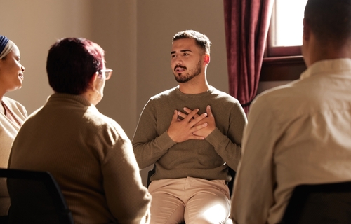 A young man holds his chest as he speaks to a small group seated in a circle.