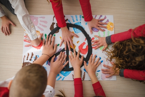 Several children add handprints to a painting with a peace symbol in the center.