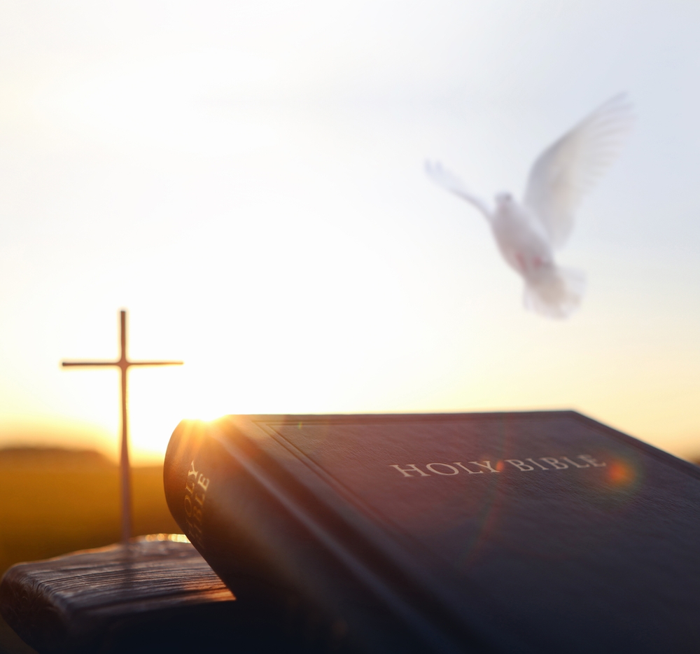 The sun sets on a Bible with a cross and a dove in the background.