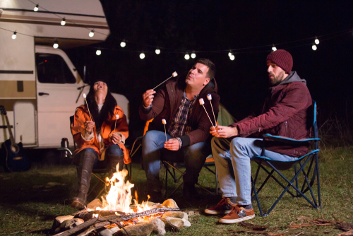Three people roast marshmallows at a campfire in front of their camper.