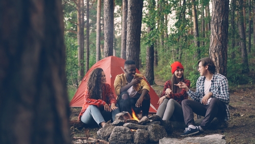 A group of young people is camping in the woods and sitting around a campfire in front of their tent.