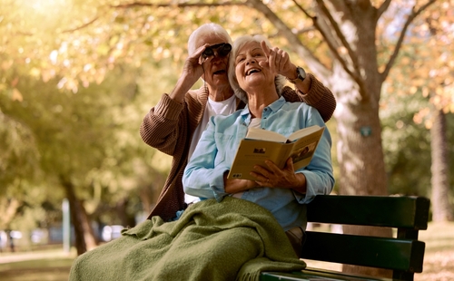 Two older people sit on a park bench and look through binoculars.