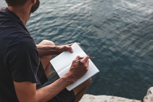A man journals in front of a body of water.