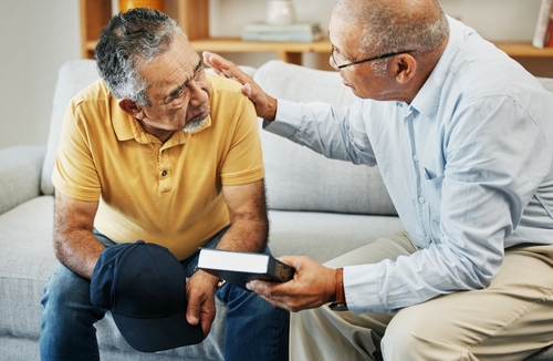 Two men sit on a couch, with one holding a Bible and patting the other one's back.