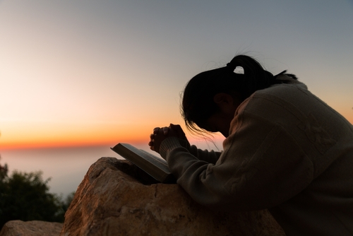 A woman prays with a Bible in front of a sunset